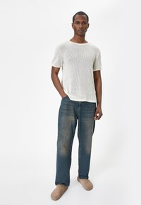 Lekki, biały sweter wykonany na drutach z krótkim rękawem, noszony z luźnymi niebieskimi dżinsami i beżowymi butami typu slip-on. Model stoi z jedną ręką w kieszeni.