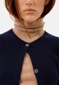 Marinblå ullcardigan med rundad halsringning, med två grönblå knappar. En beige ribbad polotröja är synlig under.