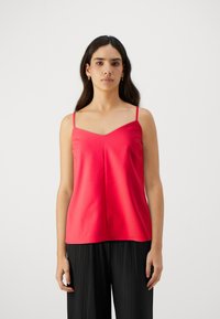 Rød cami med tynne stropper, V-hals og glatt tekstur. Bært med plissert sorte bukser, minimalistisk design, og uten synlige mønstre eller detaljer.