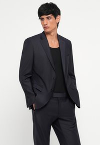 Fato azul marinho com um acabamento texturizado, apresentando um casaco de um só peito, lapelas em bico e calças tailoradas combinantes. Camiseta preta visível.