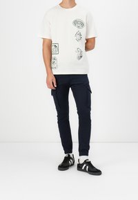 Ljusbeige T-shirt med gröna grafiska tryck; kombinerad med mörknavy cargobyxor och svarta sneakers med vita ränder. Enkelt design.