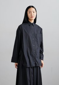Black button-up shirt with long sleeves, a chest pocket, and a loose fit. Made of smooth fabric with a slightly shiny texture.