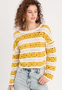Pull en crochet avec des rayures jaunes et blanches, motifs floraux et une coupe décontractée. Dispose d'un col bateau et d'une bordure côtelée.