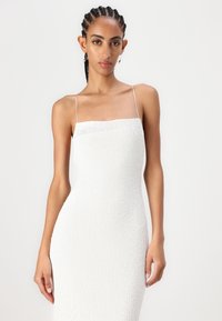 Λευκό πλεκτό φόρεμα με τετράγωνη λαιμόκοψη και λεπτές τιράντες. Το σφιχτό σχέδιο τονίζει το σχήμα του σώματος, με υφή στην επιφάνειά του.