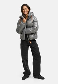 Chaqueta acolchada gris con acabado brillante, capucha desmontable y cordones naranjas. Combinada con jeans anchos negros y zapatillas negras.