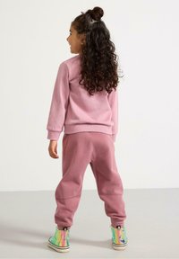 Vaaleanpunainen fleece-sweatshirt ja siihen sopivat jogger-housut, rento istuvuus. Lapsella on päällään kimaltavat korkeavartiset lenkkarit. Takaview on esillä.