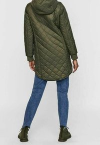 Olivově zelená prošívaná bunda s kapucí, s diagonálním vzorem a dlouhými rukávy, v kombinaci s modrými džíny a zelenými botami.