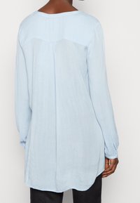 Person, der eine langärmelige, hellblaue Bluse mit lockerer Passform, gefalteten Rückendetails und geknöpften Manschetten trägt, kombiniert mit schwarzen Hosen.