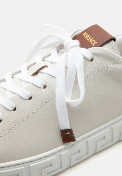 Baskets Versace en cuir blanc avec des lacets blancs noués en nœud, des accents en cuir marron et une semelle blanche à motif.