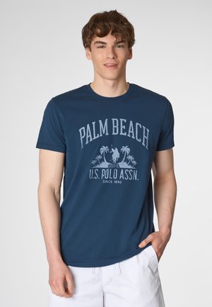 Giovane uomo con una maglietta blu navy che presenta la scritta "Palm Beach" e un logo polo, in piedi contro uno sfondo chiaro, sorridente.