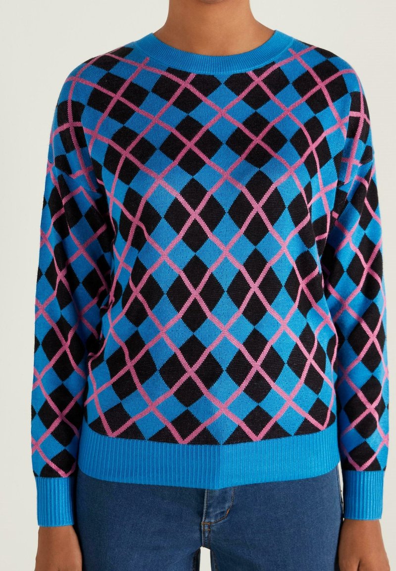 Pull bleu avec motif losanges argyle noir et rose, porté avec un jean bleu, montré du cou aux hanches.