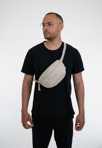 Béžová ľadvinka s hladkým povrchom, so zipsovým zapínaním a nastaviteľným popruhom, nosená cez čierne tričko na jednoduchom pozadí.