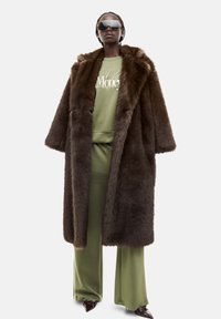 Brun faux fur frakke med en bred krave, parret med en lysegrøn sweatshirt og matchende bukser. Modellen bærer mørke solbriller og skinnende sko.