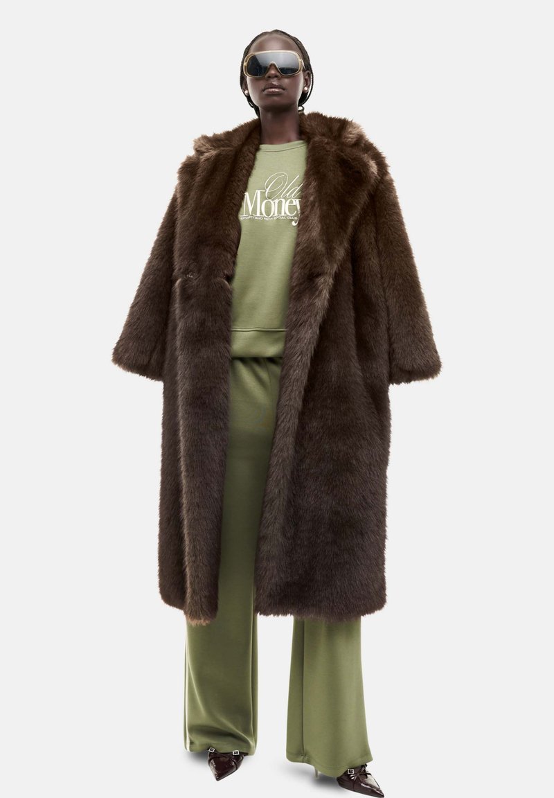 Brun faux fur frakke med en bred krave, parret med en lysegrøn sweatshirt og matchende bukser. Modellen bærer mørke solbriller og skinnende sko.
