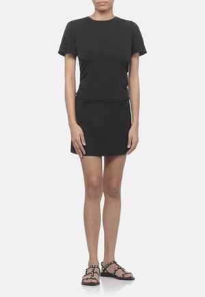 Donna in piedi che indossa una maglia nera semplice a maniche corte, una minigonna nera e sandali neri piatti con borchie su sfondo bianco.