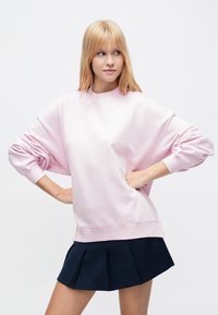 Ανοιχτό ροζ φαρδιά μπλούζα με χαλαρούς ώμους, ριμπ μανσέτες και στρογγυλή λαιμόκοψη, συνδυασμένη με μια σκούρα μπλε πλισέ φούστα.