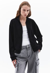 Felpa nera con zip e due tasche frontali. Presenta una vestibilità rilassata e un tessuto in misto cotone. Indossata sopra una canotta bianca e pantaloni cargo grigi.