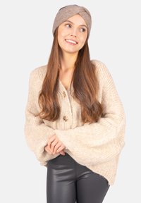 Pull en tricot beige avec des manches larges, comportant de grands boutons et une finition texturée. Assorti avec un bandeau en tricot marron et un pantalon noir.