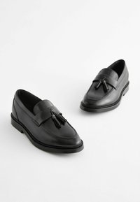 Mocassins en cuir noir à finition lisse, avec une sangle décorative et des glands, et une semelle en caoutchouc texturée et basse.