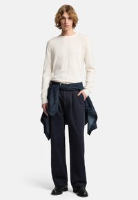 Maglione bianco a maniche lunghe, pantaloni blu scuro dal taglio comodo e una giacca di denim legata in vita. Scarpe nere completano l'outfit.