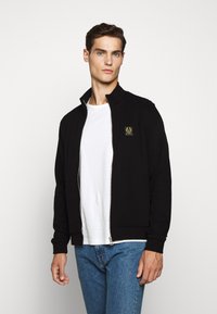 Svart zip-up jacka med ståkrage, med en liten logotyp-patch på bröstet. Buren över en vit t-shirt och blå jeans.