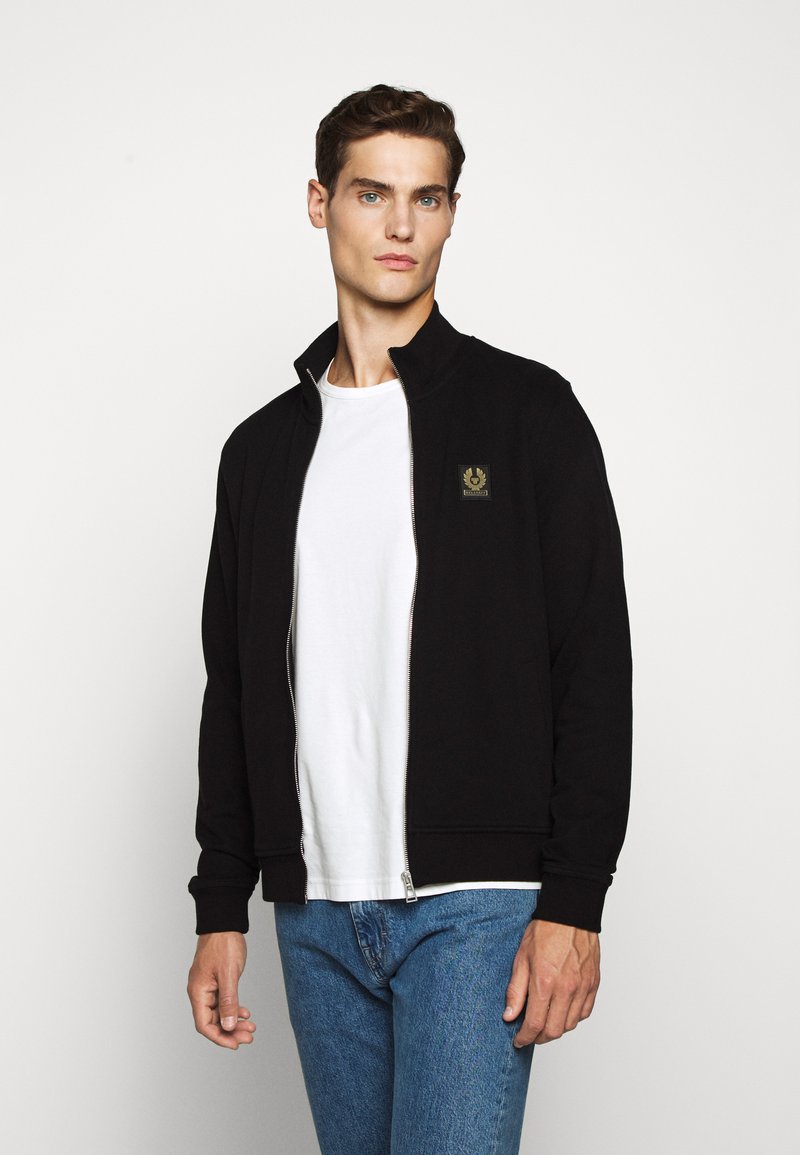 Svart zip-up jacka med ståkrage, med en liten logotyp-patch på bröstet. Buren över en vit t-shirt och blå jeans.