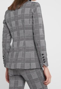 Femme vue de dos portant un costume à carreaux pied-de-poule gris et noir, avec une veste cintrée et des boutons sur la manche.