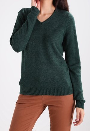 Femme portant un pull à col en V vert foncé et un pantalon marron, debout avec une main près de son visage et l'autre le long du corps.