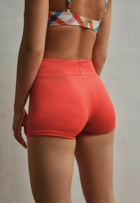 Kvinde iført koralfarvede atletikshorts med høj talje og en multifarvet ternet sports-bh, stående foran en lys, ensfarvet væg.