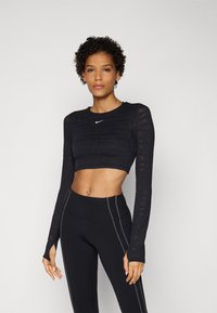 Fekete, rövidített, hosszú ujjú fitnesz felső, diszkrét mintával, kerek nyakkivágással és Nike logóval. Magas derekú fekete leggings-szel párosítva.
