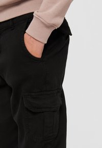 Pantalon cargo noir avec une poche latérale, associé à un pull rose clair. Le tissu semble durable avec une texture lisse.