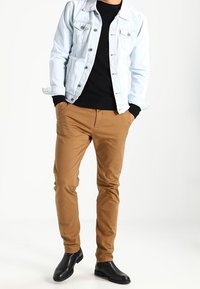 Hellblaue Jeansjacke über einem schwarzen Shirt, beige schlanke Hose und schwarze Knöchelstiefel. Die Jacke hat zwei Brusttaschen und eine durchgeknöpfte Vorderseite.