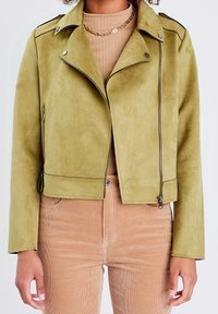 Veste de moto en faux suède vert olive avec des finitions noires, des poches zippées et une coupe courte. Associée à un haut beige avec un effet côtelé.