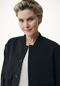 Zwarte bomberjack met een geribde kraag, sluiting met knopen aan de voorkant en twee zakken. Lichtgewicht stof met een gladde textuur, gedragen over een grijze top.