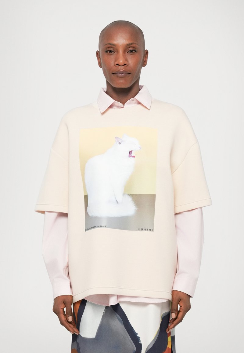 Prevelik svetlo roza pulover z belim grafičnim motivom mačke, ki ima mehak material in kratke rokave. Preliti čez svetlo roza srajco s kratkimi rokavi.