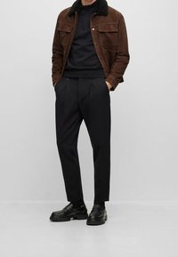 Man wearing brown jacket with black fur collar, black quarter-zip sweater, black pleated trousers, and black loafers with thick soles.