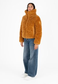 Brązowa, puszysta kurtka teddy z wysokim kołnierzem, zamkiem błyskawicznym i bocznymi kieszeniami, zestawiona z szerokimi niebieskimi dżinsami i białymi trampkami.