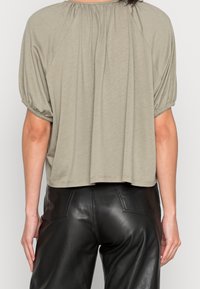 Top a maniche corte di colore verde oliva, realizzato in tessuto morbido con una vestibilità ampia e scollo arricciato, abbinato a pantaloni in ecopelle nera.