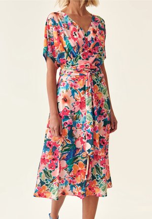 Woman wearing a colorful floral wrap dress with short sleeves and a tied waist belt, standing against a plain background.