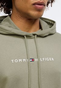 Detailný záber na osobu v svetlozelenej mikine s bielym nápisom "Tommy Hilfiger" a malým červeným, bielym a modrým logom umiestneným na hrudi.