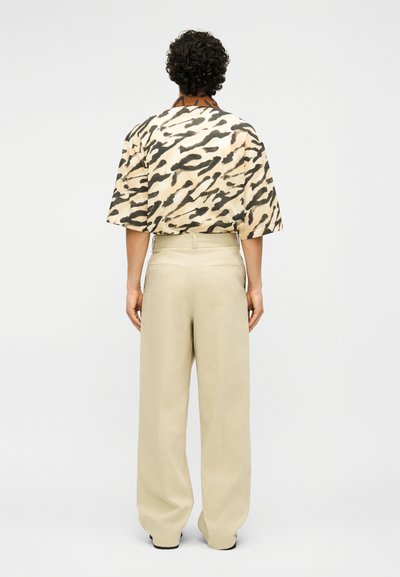 Άτομο με σγουρά μαλλιά που φοράει ένα μπεζ, μαύρο και κρεμ κοντομάνικο πουκάμισο με animal print και ψηλόμεσα φαρδιά παντελόνια κρεμ, στέκεται γυρισμένο με την πλάτη.