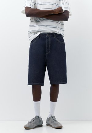 Person trägt dunkle Jeansshorts, weiße Sportsocken, graue Sneaker und ein weißes Hemd mit dünnen blauen Streifen, steht mit verschränkten Armen.