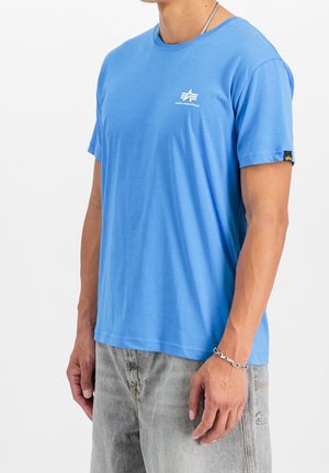 Homme portant un t-shirt bleu Alpha Industries, un collier chaîne argenté, un bracelet et un jean gris clair, debout devant un fond blanc.