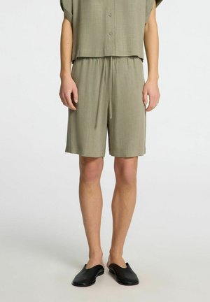 Olive green matching shorts and short-sleeve top, featuring a relaxed fit, elastic waistband, and subtle texture. Worn with black slip-on shoes.