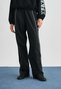 Άτομο που φοράει μαύρο αθλητικό παντελόνι με λευκές ρίγες και μαύρα παπούτσια, στέκεται σε μπλε χαλί μπροστά σε απλό φωτεινό φόντο.