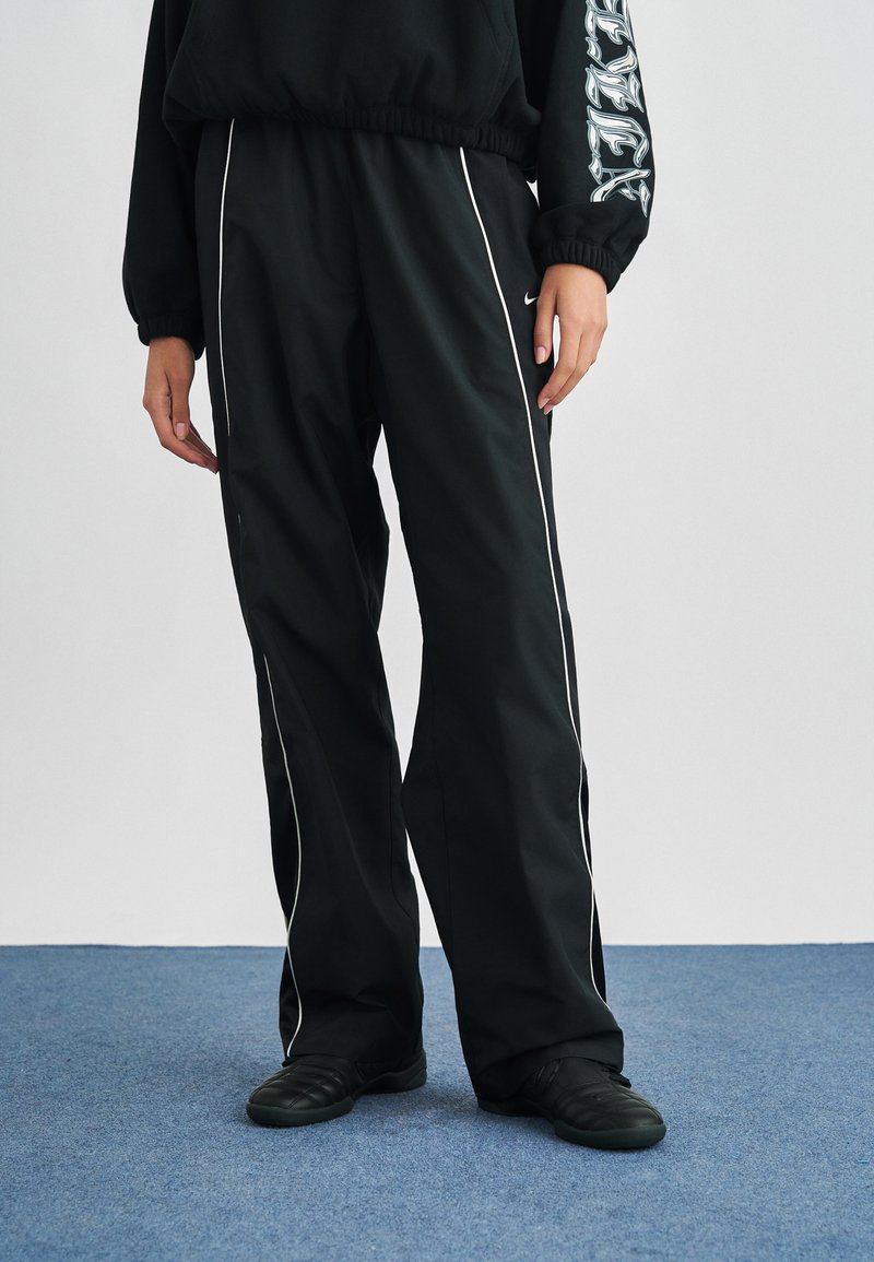 Άτομο που φοράει μαύρο αθλητικό παντελόνι με λευκές ρίγες και μαύρα παπούτσια, στέκεται σε μπλε χαλί μπροστά σε απλό φωτεινό φόντο.