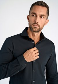 Schwarzes Longsleeve-Shirt mit spitzem Kragen, glattem Stoff, geknöpften Manschetten und subtiler Glanz. Der Träger richtet mit einer Hand den Kragen ein.