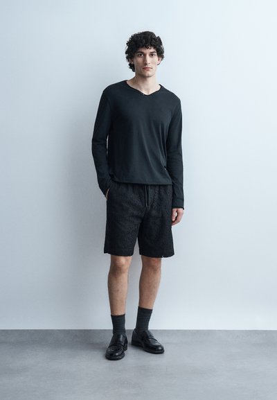 Νεαρός άνδρας με κατσαρά μαλλιά στέκεται μπροστά σε ανοιχτογρι τοίχο, φορώντας μαύρο πουκάμισο με μακριά μανίκια, μαύρα σορτς με υφή, σκούρες κάλτσες και μαύρα επίσημα παπούτσια.