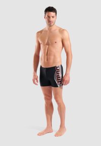 Schwarze Badeshorts mit figurbetontem Design, versehen mit einem roten und weißen Logo an der Seite und Linien, die ein dynamisches Muster erzeugen.