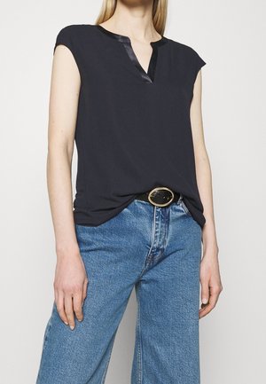 Femme portant un haut noir sans manches à col en V, rentré dans un jean bleu taille haute avec une ceinture noire ornée d'une boucle ovale dorée.